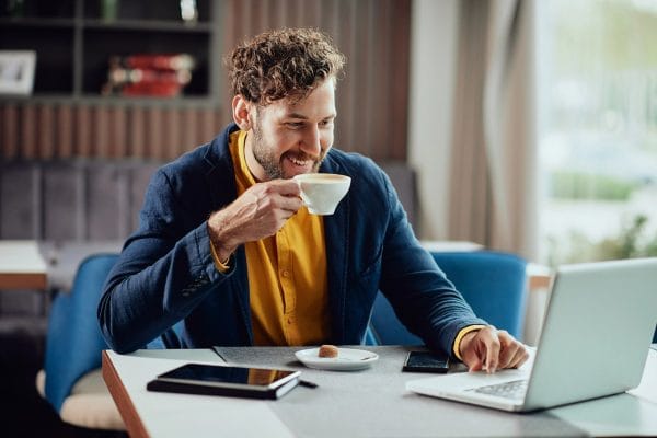 Financial planning for digital nomads and remote workers | enrizen | Close up of smiling freelancer sitting in cafe, drinking fresh coffee and using laptop