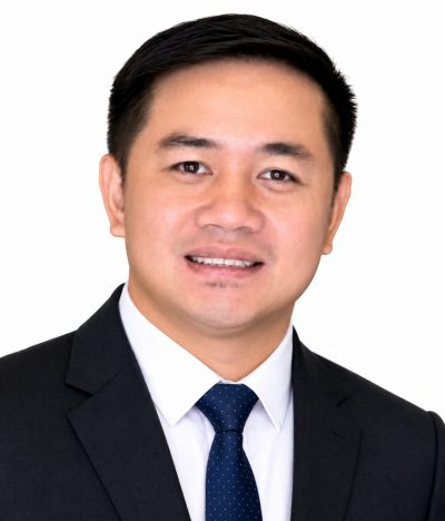 Portrait of an asian man in a dark suit, white shirt, and blue tie, smiling against a white background.